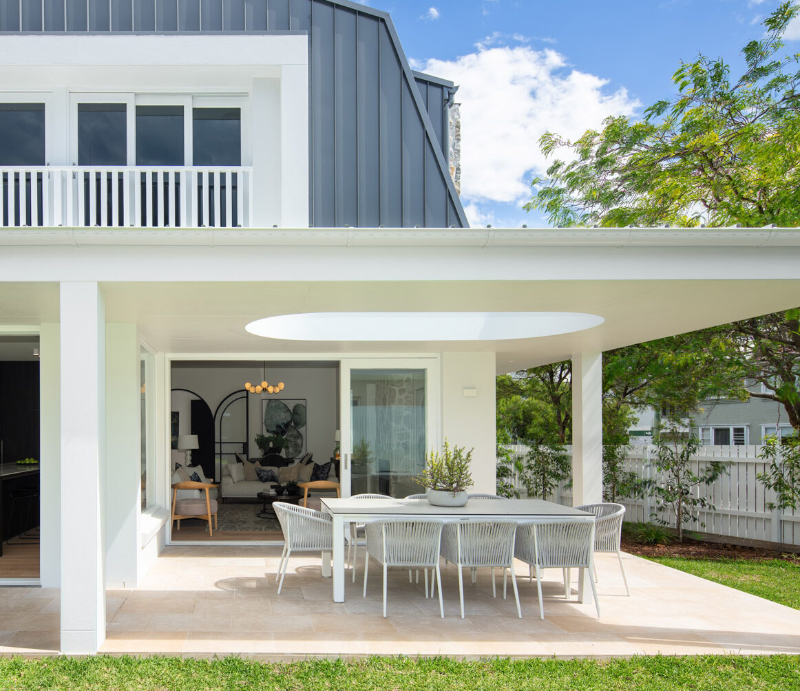 Contemporary barn-style home with white exterior, covered outdoor terrace and minimalist outdoor dining area overlooking a green lawn